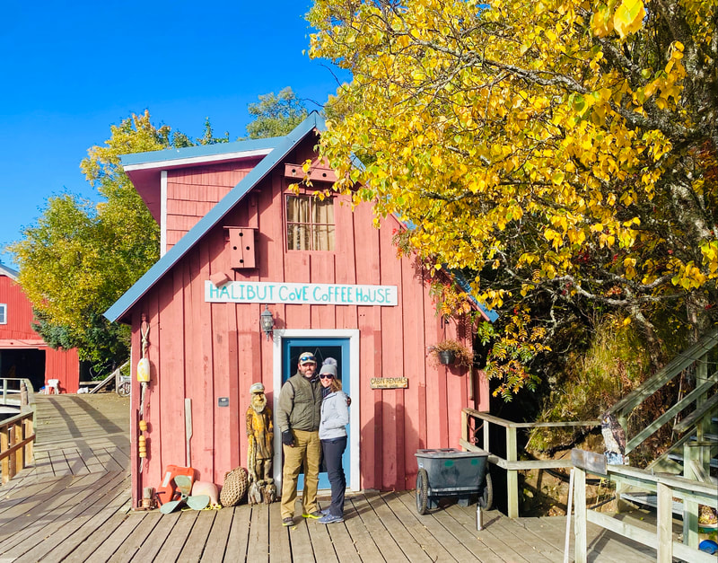 A couple posing on the boardwalk in front of the Halibut Cove Coffee House with autumn-colored trees nearby. 