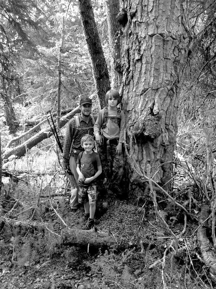 A young man posing with two kids next to a roughly textured cottonwood tree during a hike to Leisure Lake. 