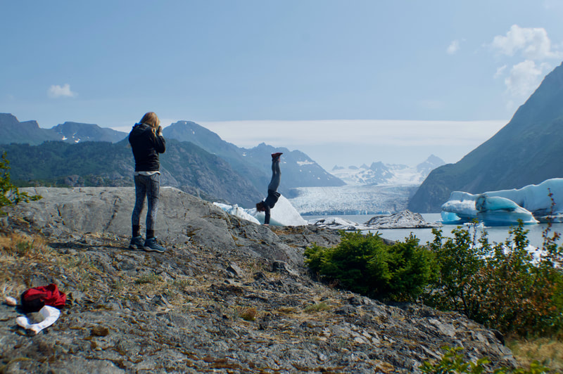 A woman taking a picture of her friend doing a handstand with Grewingk Glacier in the background. 