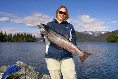 A smiling woman holding up a large King Salmon on a beautiful sunny day. 