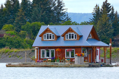 A quaint, floating house in Halibut Cove, Alaska. It has a blue, gabled roof, cedar siding, and flower baskets on the deck. 