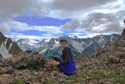 A smiling woman posing on the top of Alpine Ridge with the mountain peaks of the Kachemak Bay State Park in the background. 