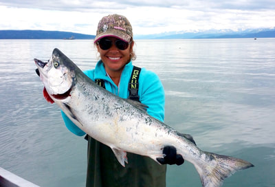 A woman holding a large salmon with Kachemak Bay and the mountains behind her. 